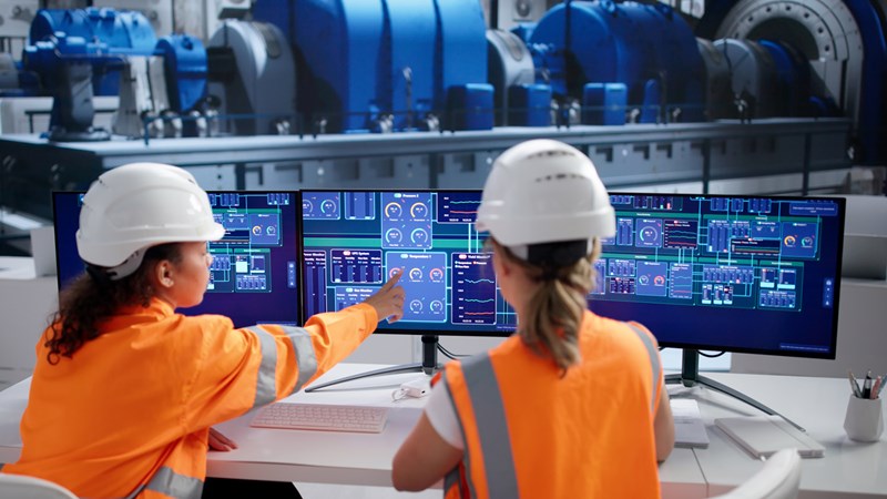 Två personer i skyddsutrustning arbetar vid flera stora bildskärmar som visar tekniska processer i en industriell kontrollmiljö, med maskiner synliga i bakgrunden.