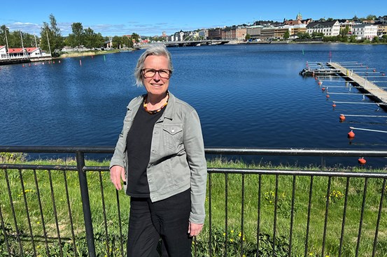Christina står vid räcke vid vatten i stadsmiljö med bro, byggnader och småbåtshamn i bakgrunden, soligt väder.