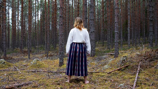 Foto. En kvinna står barfota mitt i en tallskog med ryggen mot betraktaren. Hon har blont hår, vit blus och mörkt blå kjol med röda och vita inslag.