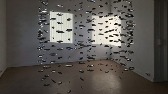 I ett rum med dämpat ljus syns en installation som består av spetsiga ovaler som hänger från taket i en spiral. Installationen påminner om ett fiskstim och blänker i solljuset från fönstren.