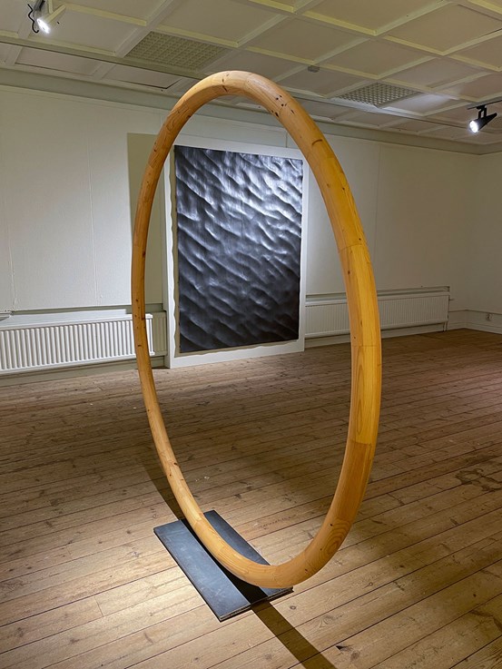På ett ljust trägolv står en stor ring av trä snett vinklad mot betraktaren. Som fot har den en mörk metallplatta. I bakgrunden syns en stor mörk bild på en vägg.