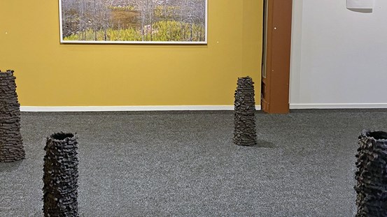 På en grå heltäckningsmatta står gråsvarta tornliknande skulpturer. I bakgrunden på en gul vägg skymtar en landskapsbild. 