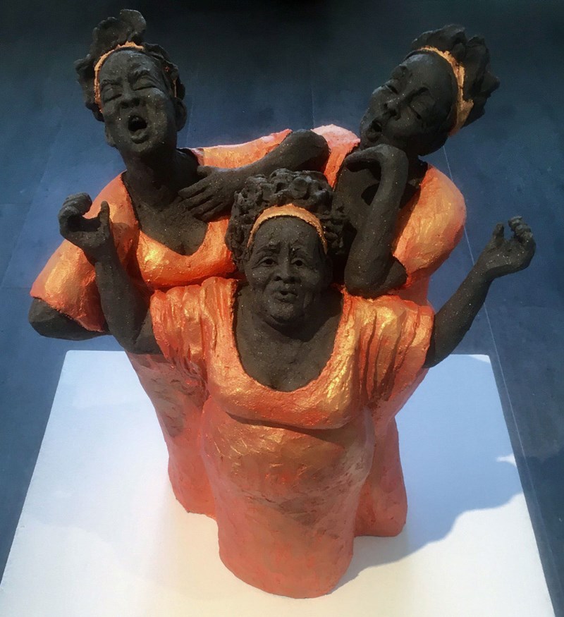 På ett vitt podium står en skulpur, en kör bestående av tre kvinnor i mörk keramik. De har röd-orangea blanka klänningar och matchande hårband. Alla har öppen mun och ser ut att sjunga inlevelsefullt. 