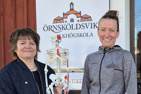 Anna Viström och Elena Dahlén håller ett litet skelett mellan sig där de står framför en skylt med folkhögskolas logga. 