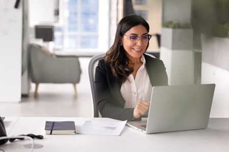 Mörkhårig kvinna med glasögon som sitter vid dator i hemmiljö och tittar på datorskärmen.