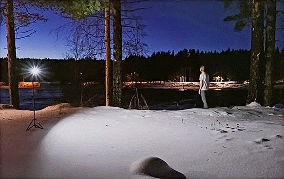 Foto, en landskapsvy på kvällen med den snöiga förgrunden upplyst av en lampa på stativ till vänster i bild. En person står bortvänd och tittar ut över mörkt landskap och djupblå himmel. 