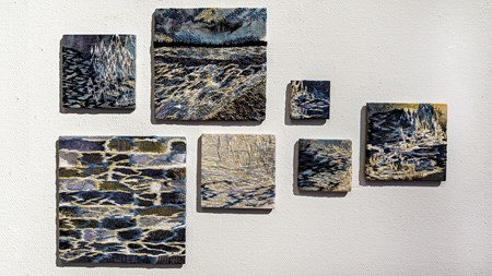 Sju kvadaratiska verk i olika storlekar på en vit vägg. Ett fruset landskap i vita, gula, blå nyanser i skarpa kontraster; iskallt strömmande vatten, vassruggar, mörk skog och isande vind.