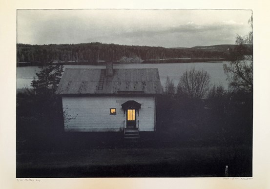 Landskap i skymning, ett vitt litet hus. Gult ljus från dörrens fönster och det intill. Bortom, ett vattendrag. Blek himmel. 