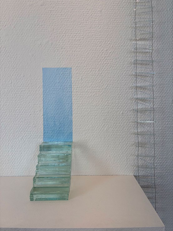 En glastrappa leder upp till en rektangulär ljusblå yta målad på väggen. Till höger en stege av glas och ståltråd. 