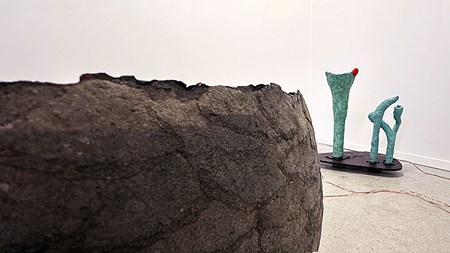 I förgrunden syns en del av en stor, rund form, brun med skrovlig yta. I bakgrunden några gröna, avlånga skulpturer. 