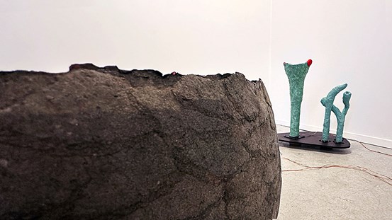 I förgrunden syns en del av en stor, rund form, brun med skrovlig yta. I bakgrunden några gröna, avlånga skulpturer.