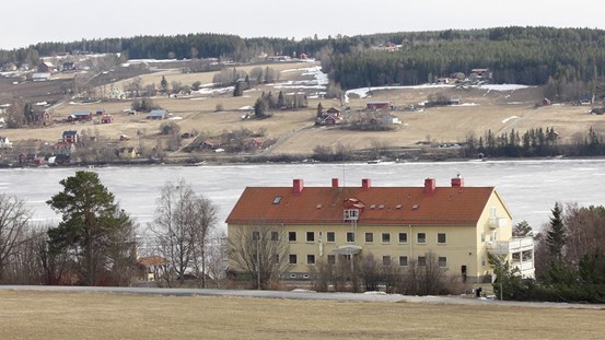 En beige stor byggnad vid vatten, på andra sidan älven syns hus, öppna fält och skog. 