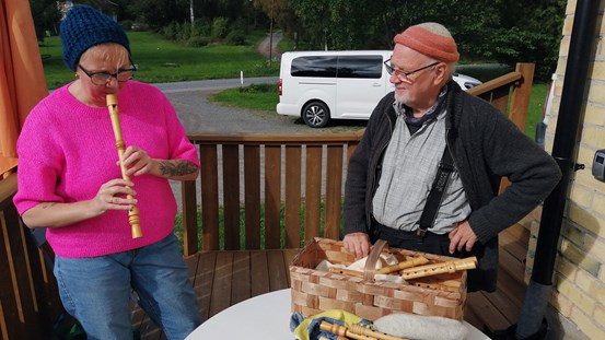 En kvinna och en man står utomhus på vad som kan vara en veranda. I bakgrunden syns gröna träd, en väg och en parkerad vit bil. Kvinnan till vänster har glasögon, en blå mössa, rosa stickad tröja och blå jeans. Hon håller en flöjt mot munnen och det ser ut som hon spelar på den. Mannen, till höger, är vänd mot kvinnan och tittar på. Han har lite grått skägg, glasögon och en rödorange mössa högt på huvudet, en grå skjorta och hängslen under sin mörka jacka. På bordet framför honom står en näverkorg med fler flöjtar i. 