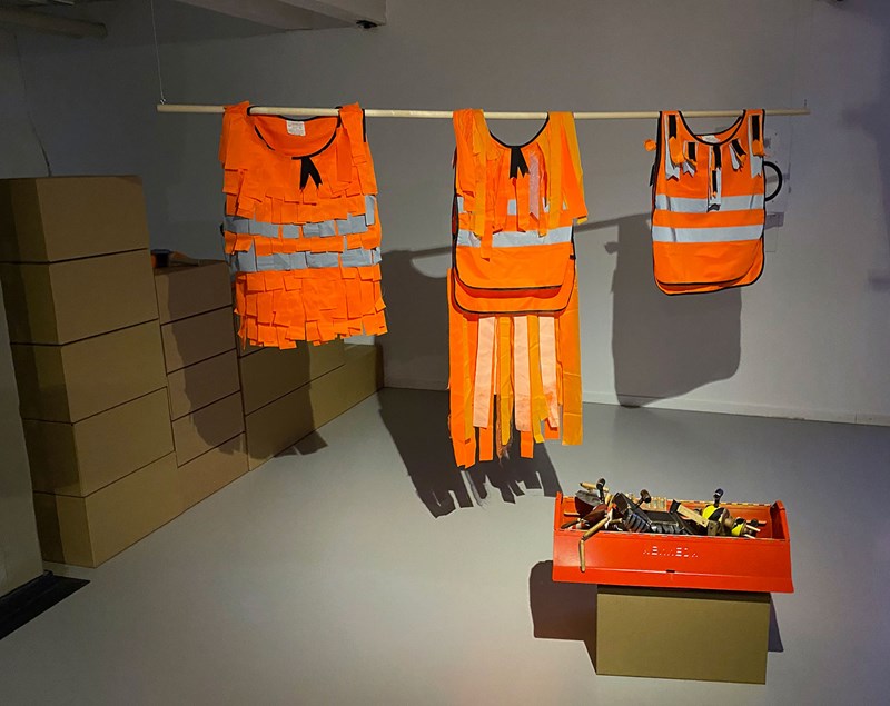 Från taket i ett vitt, kalt rum hänger en träribba. På träribban hänger tre orangefärgade varningsvästar med gråa reflexränder på. De är delvis uppklippta i remsor och ser även ut som fler remsor är ditsatta. Framför västarna står på golvet en låda med saker i. Till vänster i bakgrunden syns flera bruna papplådor travade på varandra.