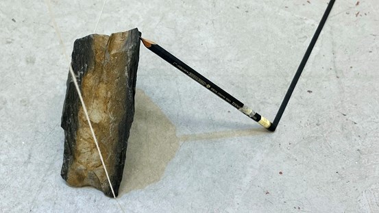 På ett gråspräckligt golv står till vänster i bild en svart och brun sten, en svart penna lutar från höger med udden mot stenen och andra änden i golvet. Därifrån löper ett svart streck, antagligen svart tejp, diagonalt uppåt höger. Från stenens bas går spända ljusa trådar.