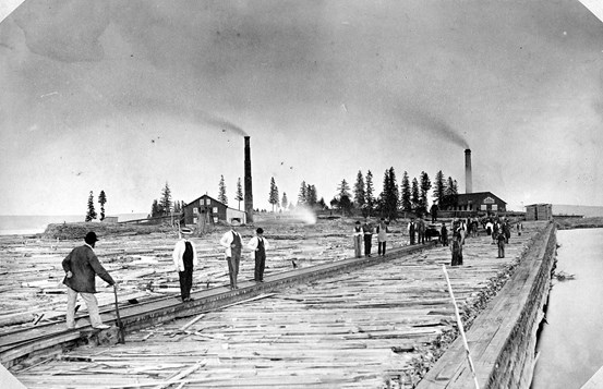 Svartvitt äldre foto från industriepoken i Timrå runt år 1870. Människor och skorstenar syns i bilden.