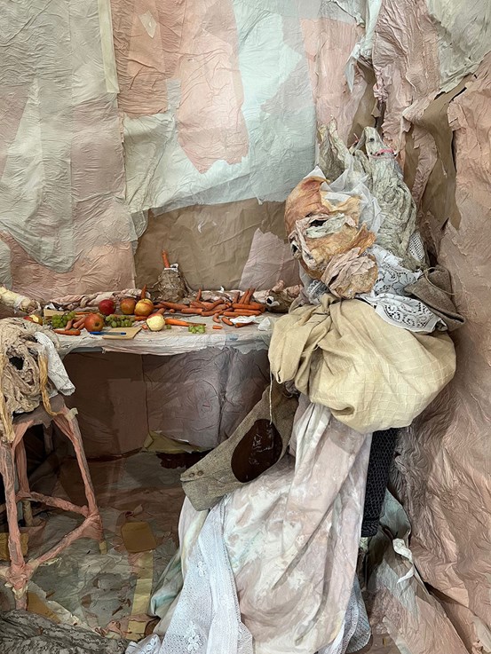 Ett rum i hopfogat av papperssjok. På ett bord ligger saker utspridda, bl.a. frukter och grönsaker. En figur av papper sitter vid bordet. 