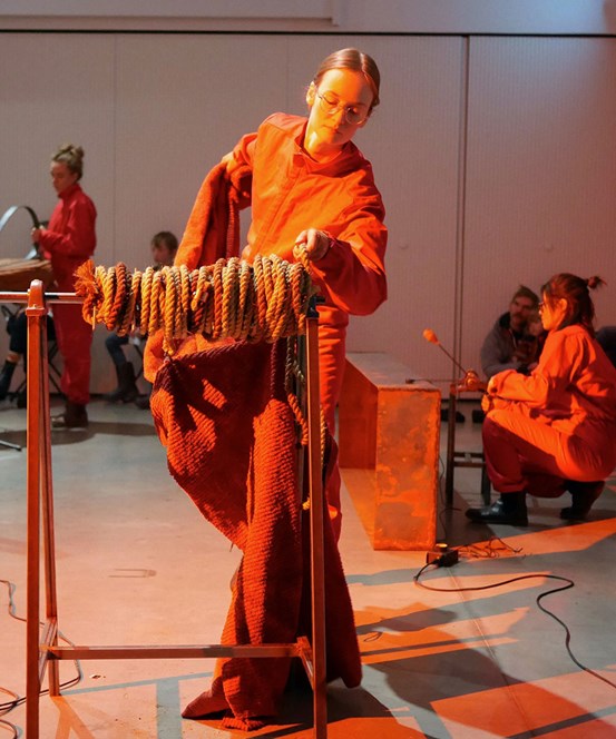En person i orange kläder står vid en träställening med ett rep uppsnurrat på. I bakgrunden syns andra personer i orange overaller upptagna med var sin sak. 