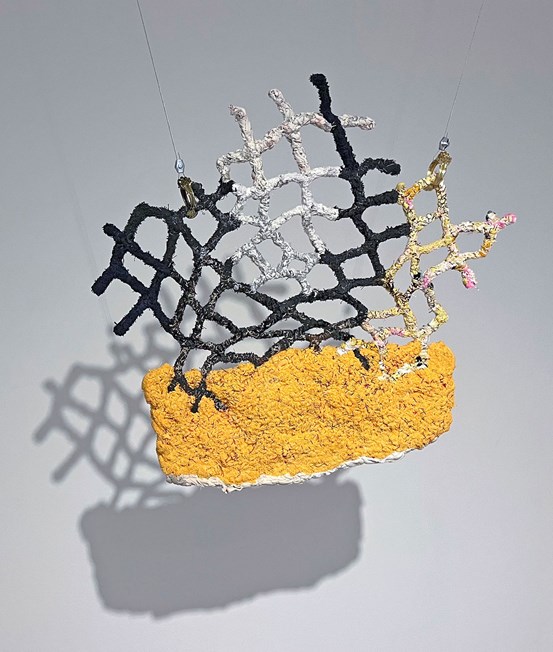 En textil skulptur hänger från taket. Ett grovt, lite spretigt nät i svart, vitt och gult sitter ihop med en gul yta under. Objektet kastar en fin skugga på väggen bakom.