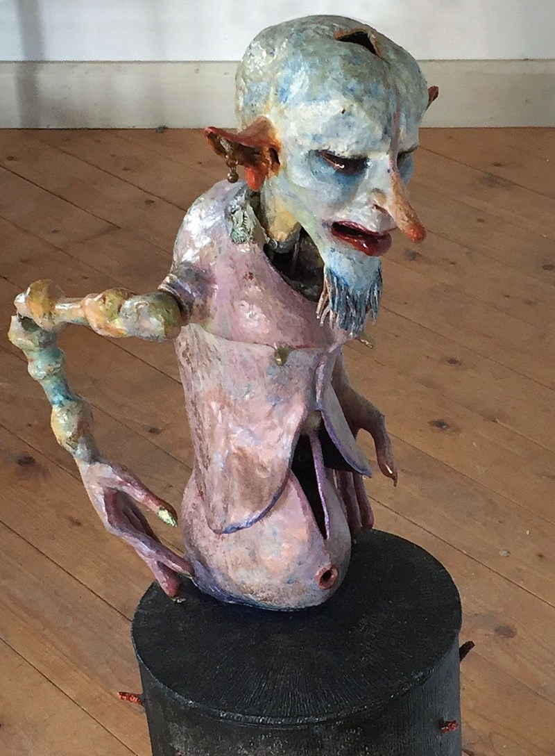 En skulptur av en skallig figur med lång vass näsa,  kisande blick, slipprig mun, spretiga fingrar med långa naglar  och tunn överkropp med kulmage. 