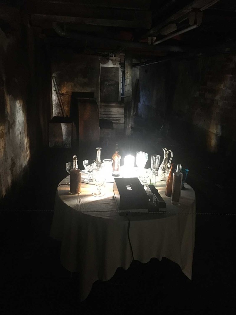 En ljusinstallation. Ett bord, glas, karaff, kristall, som konstnären hittat på området, ljus som snurrar kring bordet och skapar ett vackert skådespel som kontrasterar med den råa och underjordiska tillvaron. 