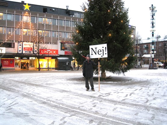 Ett foto av en man på ett torg i en stad. Han står framför en stor gran med belysning i. Det är ett tunt lager snö på marken. Han har mörka kläder och en mössa. I den en handskbeklädda handen håller han en skylt med texten ”Nej!” på. 