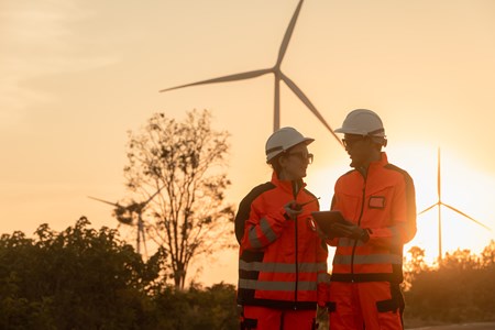 Vindkrafttekniker ute på fält, i bakgrunden syns ett vindkraftverk. 