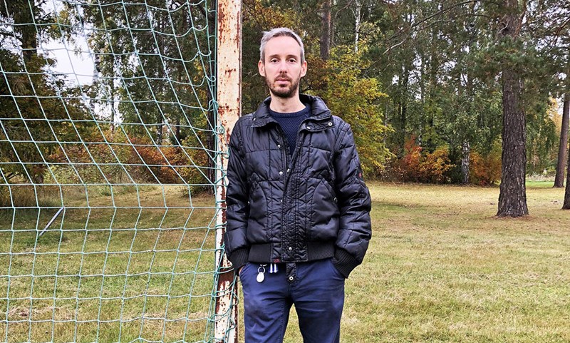 Foto av Alessandro Perini. En man med blå byxor och svart jacka står vid stolpen på ett fotbollsmål. Han har kort gråsvart hår och mörkt kort skägg. I bakgrunden syns träd och buskar i höstfärger. 