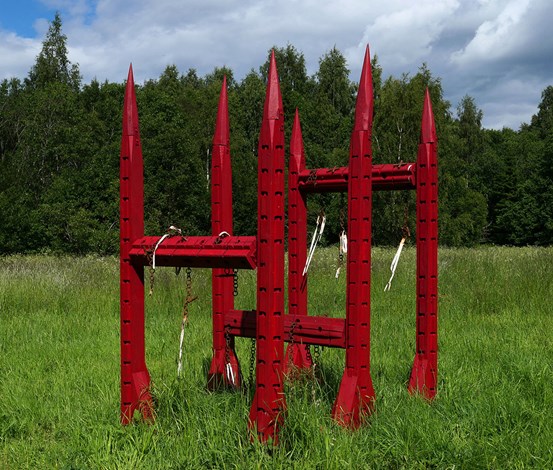 Mitt i det gröna långa gräset med lövskog som bakgrund står en skulptur. Den är gjord i trä som målats rött, med spetsiga ”torn”. Kedjor och tygstycken hänger från den. 