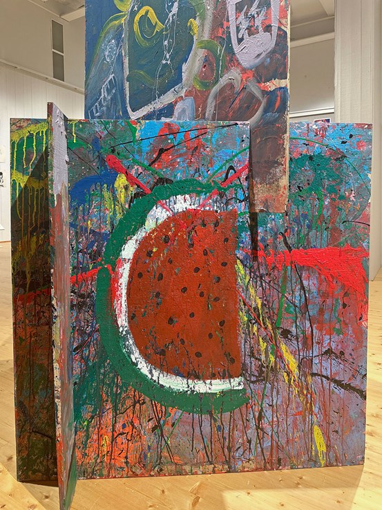 Flera träskivor med stänk, rinningar, mönster, fläckar i alla möjliga färger. I förgrunden syns en halv melon målad. 