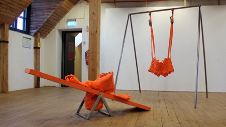 Ett rum med trägolv, vita väggar med en gungbräda i orange med orange textil på och gungställning med en orange gunga i tyg. 