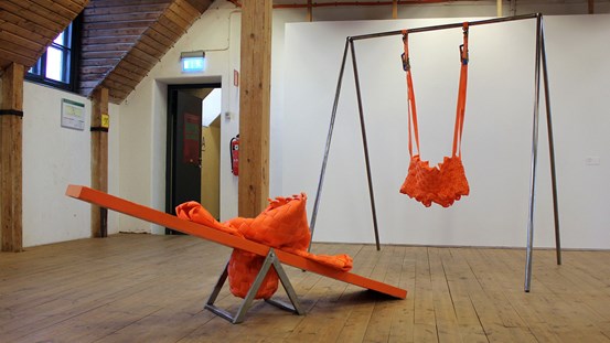 Ett rum med trägolv, vita väggar med en gungbräda i orange med orange textil på och gungställning med en orange gunga i tyg.