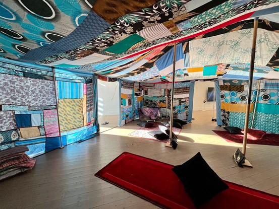 Foto: Inuti ett stort nomadtält i konsthallen, det är sytt av återvunna kläder. På golvet ligger några mattor och kuddar. 