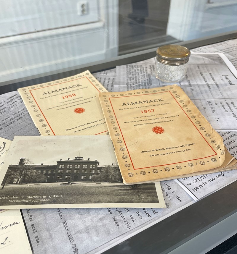 Vy över innhållet i en glasmonter. Ovanpå flera A4-sidor med text ligger två almanackor från 1955 och 1958, ett foto i svartvitt på en byggnad och en rund glasburk med ett metallock. 