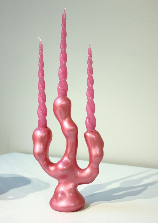 Foto: På en vit yta och med vit bakgrund står en skulptur i form av en rosa trearmad kandelaber. Formen är mjuk och bubblig och ljusen är rosa och spiralformade. 