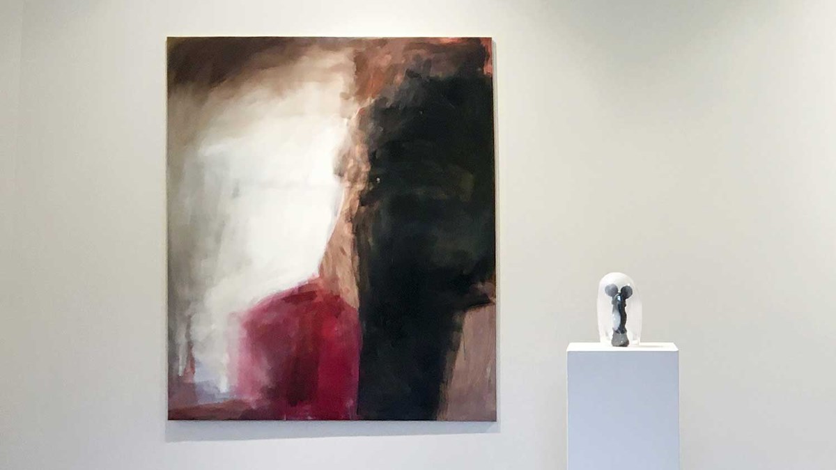 alt='På väggen hänger en målning med olja på duk, storskalig och abstrakt, med varma toner i rött, brunt, svart och vitt. På ett podium står en glasskulptur i form av en immig glaskupa med en svart seriefigursliknande form stående på insidan. '