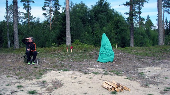 Foto av Håkan Brings performance. En gråhårig man i svarta kläder och gröna gymnastikskor sitter på en stolsryggsäck till vänster i bild. Han är tittar mot vänster i bild och håller högerhanden skuggande ovan sina glasögon. Det står en termos på marken bredvid. Där ligger också en lång remsa textil som går runt i en spiral. I mitten av bilden och spiralen står ett objekt invirat i något som ser ut som turkosblå presenning. Bakom står några tallar och längre bort fler träd. Marken är delvis sand.