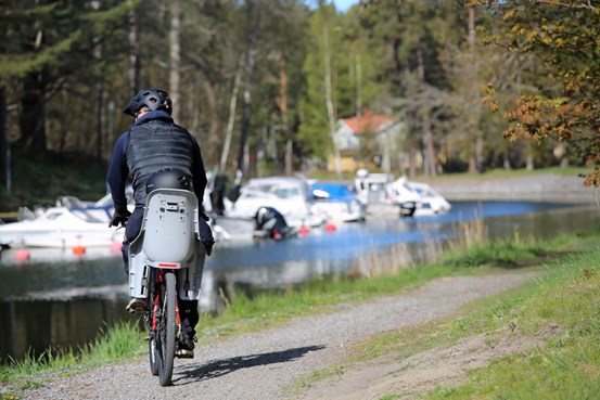 Cyklist med cykelbarnstol på cykeln, vid ett vattendrag med båtar synliga i bakgrunden.