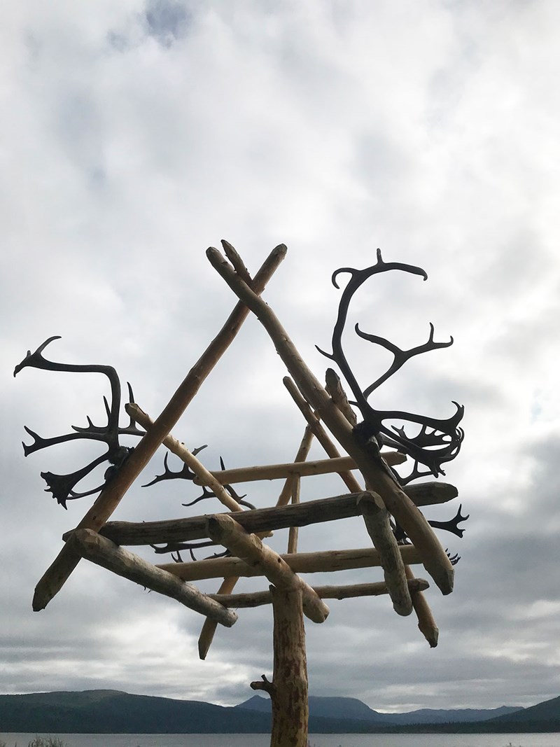 En golv- och taklös träkonstruktion av kala grenar och älghorn, dokumenterad underifrån mot en grå himmel. 