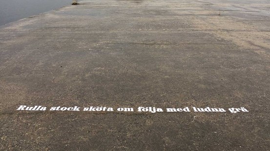 På kajens betongplatta, skrivet med stora vita bokstäver, kan man läsa första versen av konstnärens sonettkrans ”Rulla stock sköta om följa med ludna grå”.