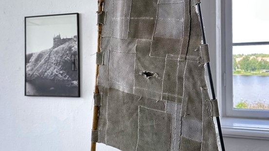 En detalj av ett verk. En del av en lappad beige-brun textil som ser ut att vara kanvas. Den är fast satt med hällor på en träpinne på ena sidan och en av metall på andra sidan. I bakgrunden till vänster på en vit vägg hänger ett foto. Till höger i bild syns en del av ett fönster i konsthallen med utsikt över vatten. 