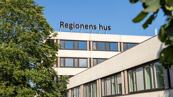 Foto av en del av en byggnad, ett grönskande träd till vänster och några blad från ett annat träd syns suddigt till höger. Längst upp på byggnaden syns en stor skylt med texten Regionens hus. I bakgrunden en blå himmel.