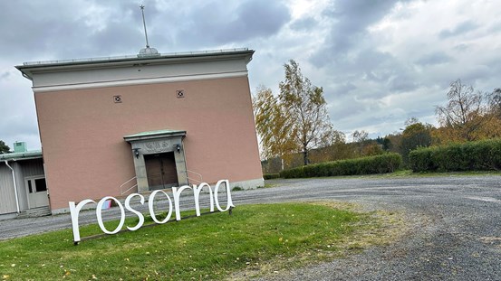 Framför en rosa byggnad är en grusväg och en rondell. Mitt i den gräsbeklädda rondellen finns ordet ”rosorna” uppmonterat. 