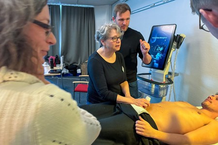 Kursdeltagare för ultraljudsapparat mot buken till mann som ligger med bar överkropp på en säng. Fler kursdeltagare står runt omkring och tittar på en skärm.