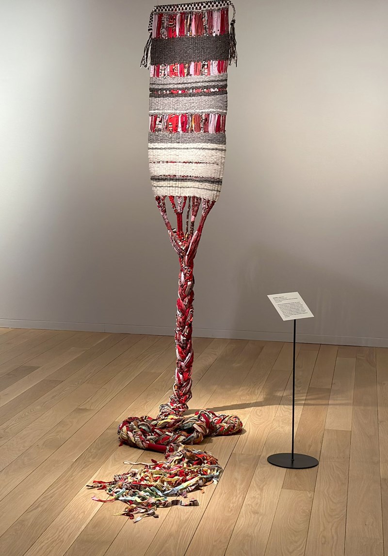 Ett skulpturalt verk i textil. Vävar och textil hänger från taket och övergår till en fläta som ringlar sig ut på trägolvet. Allt i röda, vita och grå toner. 