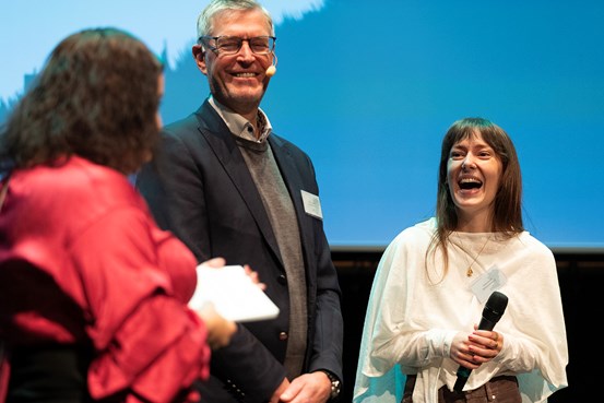 Deltagare på scen under ett seminarium. En person håller en handmikrofon, en annan använder headsetmikrofon.