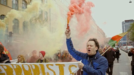 Stillbild ur video. Demonstrationståg på stadsgata. I förgrunden en man i blå jack som håller i en banderoll med en hand och i den högra uppstäckta handen en behållare som sprutar ut röd rök. I bakgrunden syns fler personer, gul rök och byggnader. 