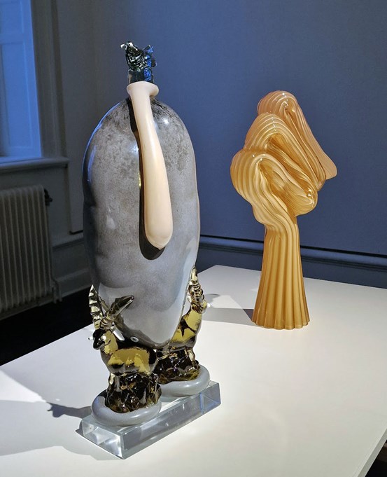 På en vit yta står två skulpturer. En skulptur i glas, en blå fågel högst upp, balanserande på en blåst form som liknar en kondom. Den ljust aprikosa, kondomliknande formen rinner ner över en stor, gråmelerad blåst glaskropp, som i sin tur vilar mellan hornen på ett par blanka antiloper. De formgjutna antiloperna vilar på varsin grå kudde, som slutligen är monterade på en kristallklar, rektangulär sockel. En skulptur i beige-orange färg står snett bakom. Den är som en utdragen rund strut där den övre delen har vikts flera gånger. Hela skulpturen har en veckig form.