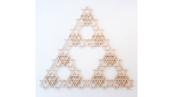 På vit vägg sitter många mindre trianglar av trä i form av en stor triangel. Tillsammans formar de mönster, större trianglar