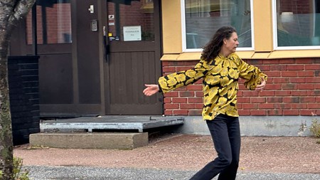 En kvinna med mörkt långt hår, gulblommig blus och mörkblå byxor dansar framför en tegelbyggnad. På dörren till byggnaden finns en skylt, ”Personalrum” står det. 